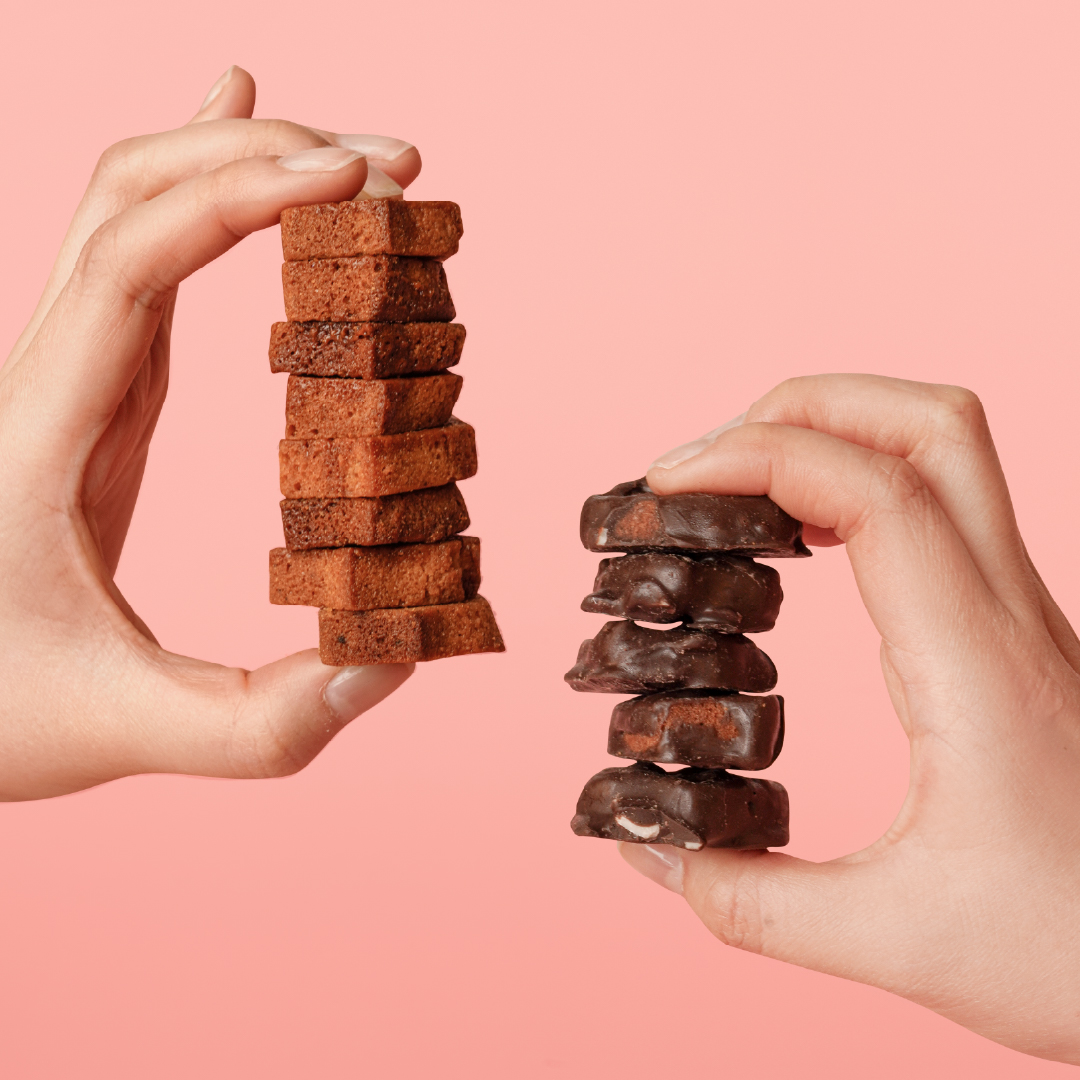 Hands holding stacks of chocolate-coated and plain financier rusks against a pink background